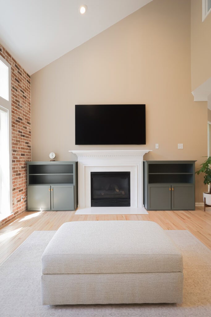 modern living room with a white fireplace and a black flat screen tv above it, flanked by two green built in cabinets. there is a beige ottoman in the foreground and a brick accent wall on the left.