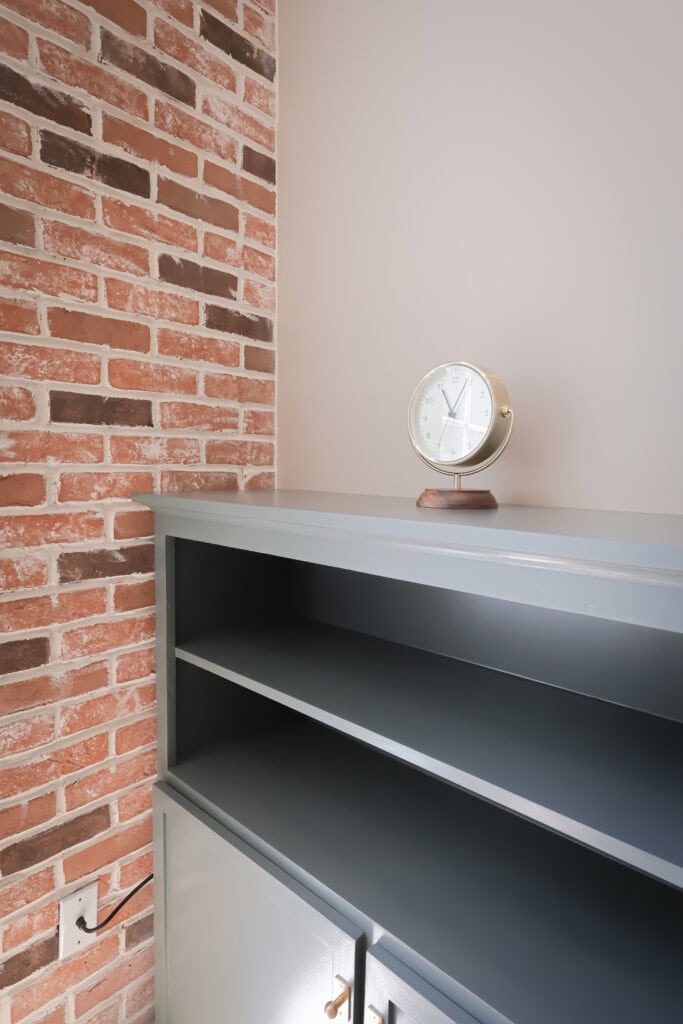 a small round clock with a gold frame sits on top of a gray shelf against a brick and beige wall. the shelf has open compartments and closed cabinets below. an electrical outlet is visible on the brick wall.