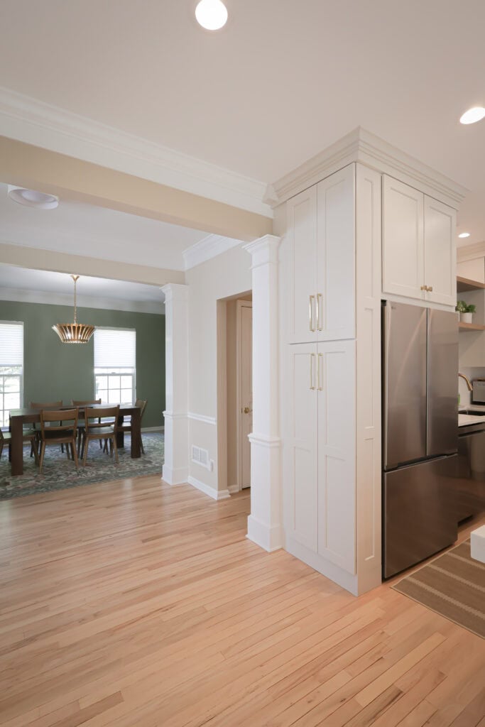 modern kitchen with light wood floors, white cabinetry surrounding a stainless steel refrigerator, and a view into a dining room with green walls, large windows, and a wooden table with chairs under a pendant light.