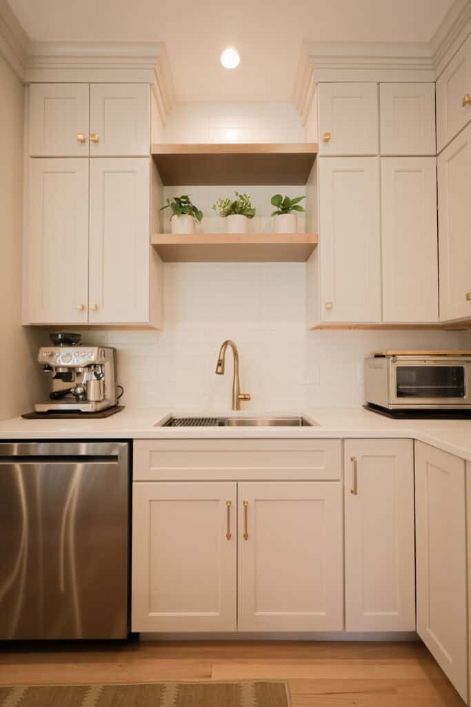 a modern kitchen features white cabinets, a stainless steel dishwasher, a sink with a gold faucet, a coffee machine, a toaster oven, and potted plants on open shelves above the sink.