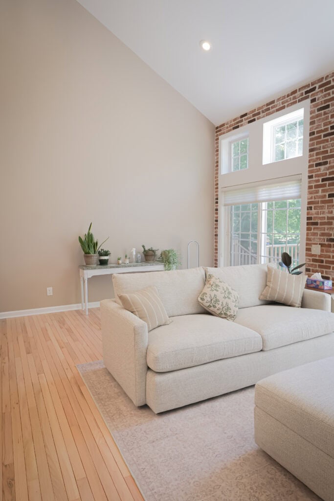 a bright living room with light wood floors, a cream sofa with patterned pillows, a beige rug, a plant filled console table, and brick accent walls with large windows letting in natural light.