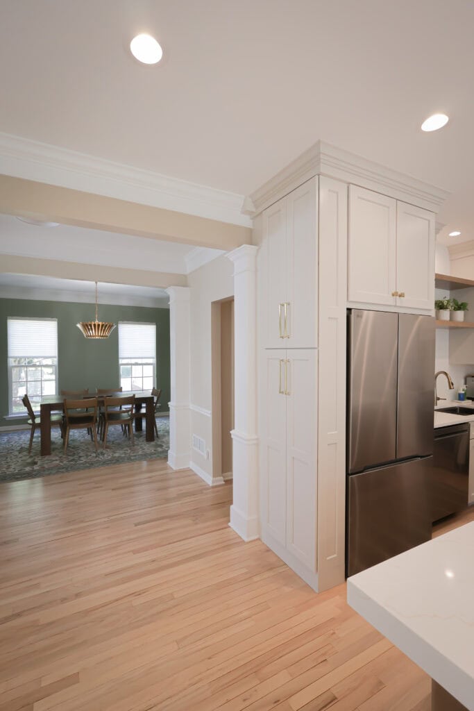 modern kitchen with light wood floors, white cabinets, and stainless steel appliances. in the background, there is a dining room with a dark table, chairs, a rug, and a gold chandelier. natural light enters through large windows.