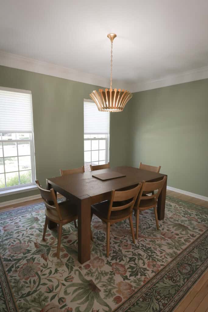 a dining room with a wooden table and six matching chairs sits on a floral rug. two large windows let in natural light, and a modern chandelier hangs from the ceiling. the walls are painted a soft green.
