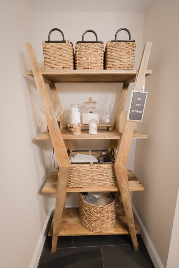 a wooden shelving unit in a corner holds wicker baskets, jars, and toiletries. a small sign reads do not disturb. the shelves display organized and neatly arranged household items.