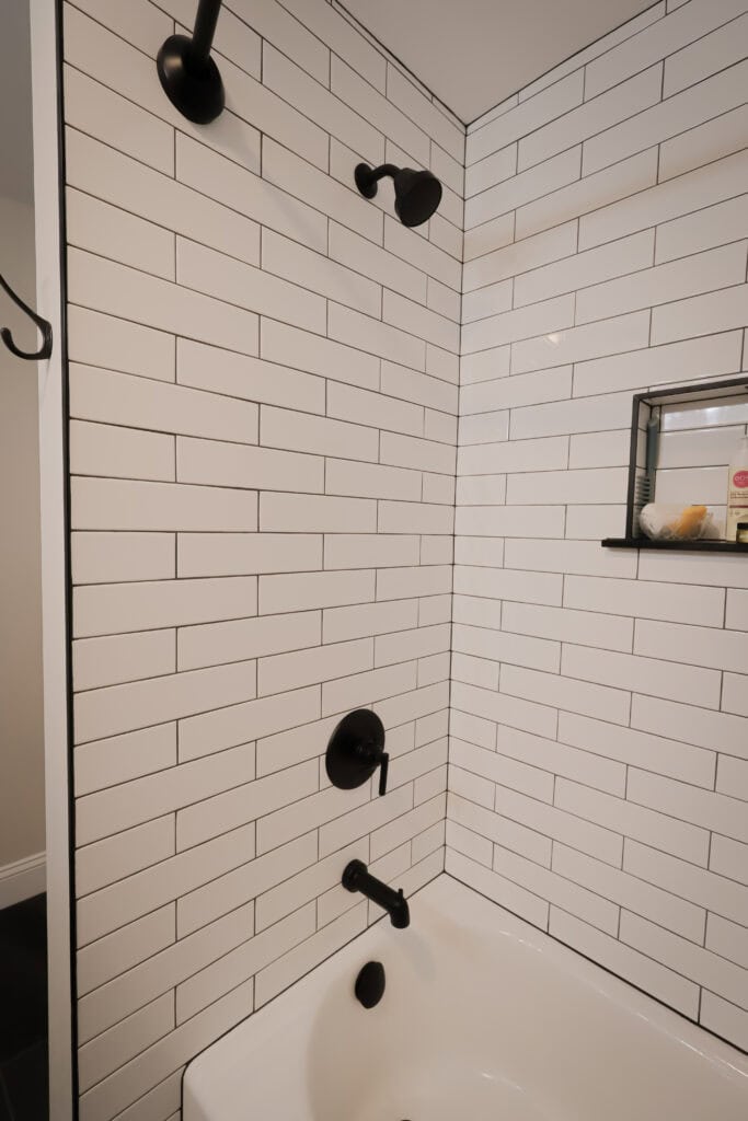 white subway tile shower with black fixtures, including a showerhead, faucet, and handle. built in niche with toiletries is visible. the tub is white and the overall style is modern and minimalistic.