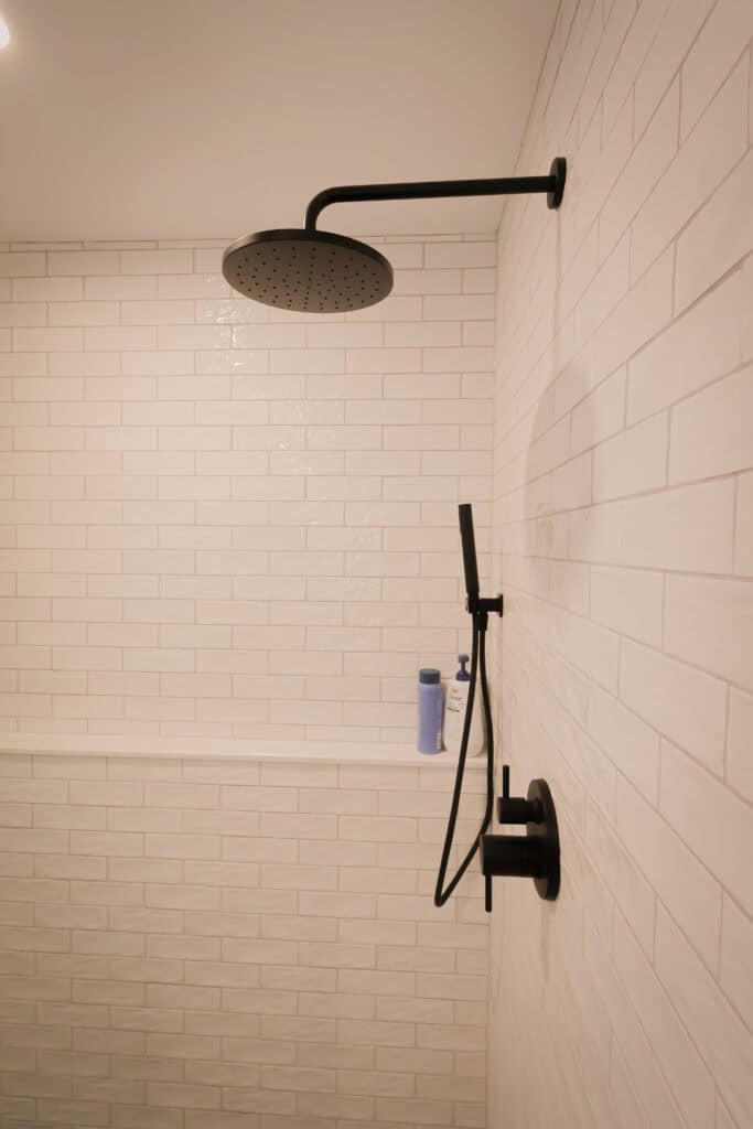 A modern shower with white subway tile walls, a matte black rain showerhead, matching handheld shower, and a built-in shelf holding a bottle of shampoo and a blue item.