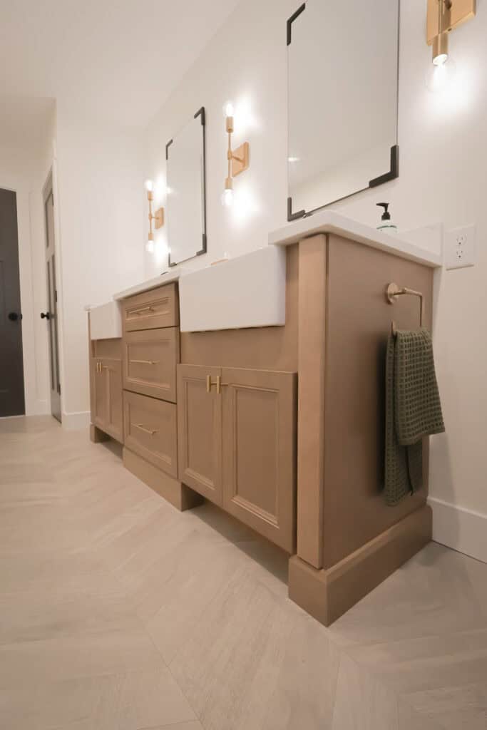 Modern bathroom with two rectangular mirrors, double sinks, wooden cabinets with gold handles, a green towel hanging on the right, and light fixtures mounted above the sinks. The floor has light beige tiles in a herringbone pattern.