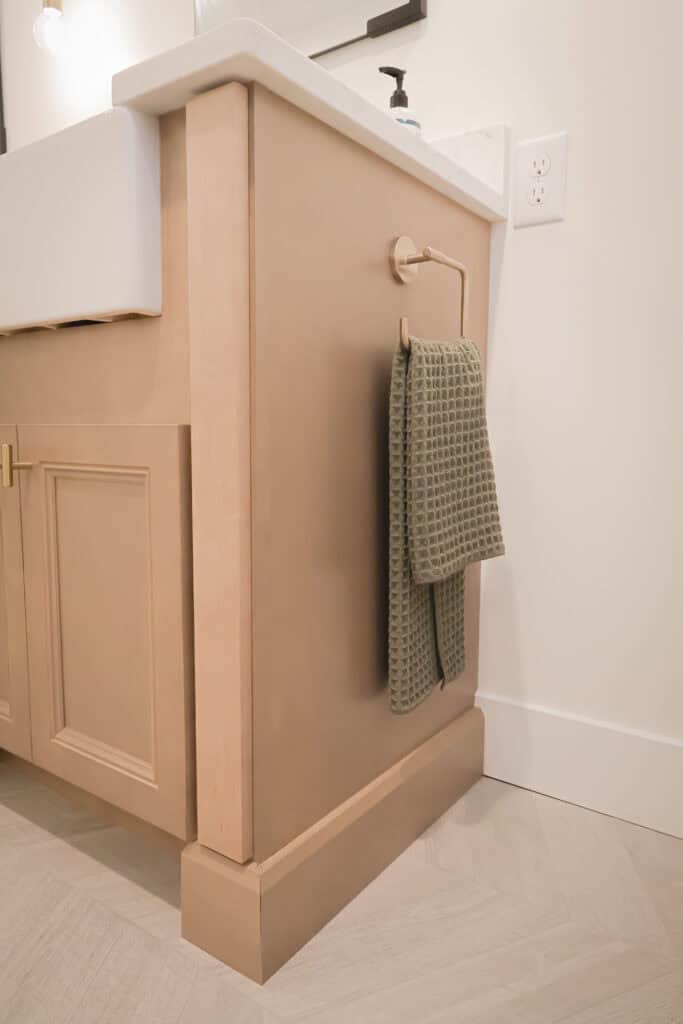 A beige bathroom vanity with a green textured hand towel hanging on a gold towel ring attached to the side. The background includes a white wall, electrical outlet, and light-colored herringbone floor tiles.