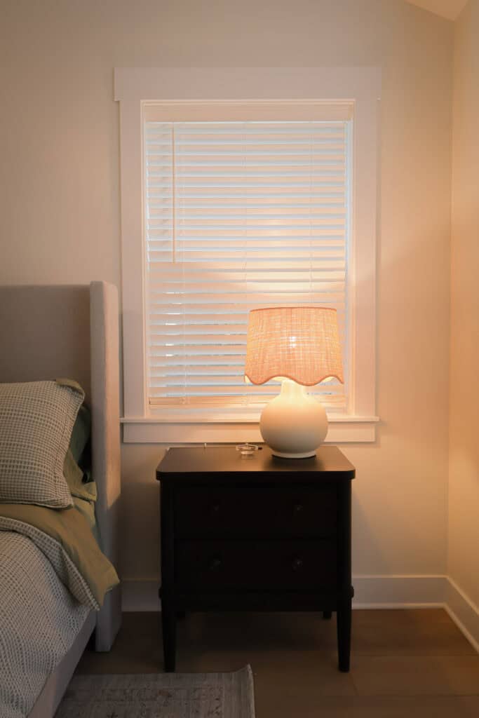 A bedside table with two drawers holds a white lamp with a woven shade, next to a neatly made bed with green and gray bedding. A window with white blinds is above the table, letting in soft light.