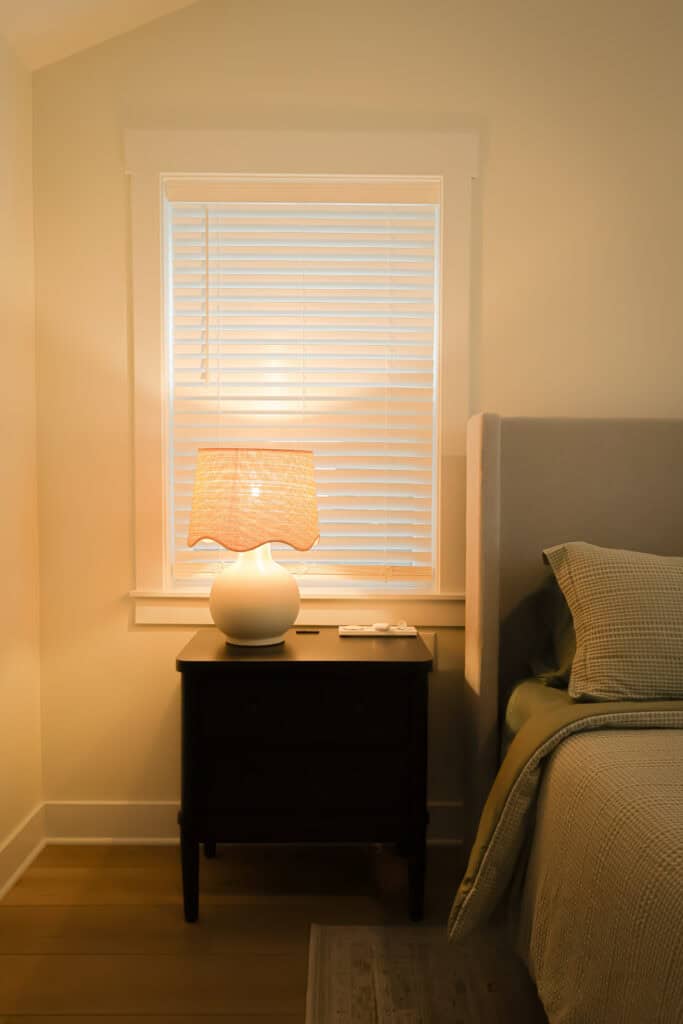 A bedside table with a lit lamp, remote controls, and a window with closed blinds beside a neatly made bed with green and beige bedding in a softly lit bedroom.