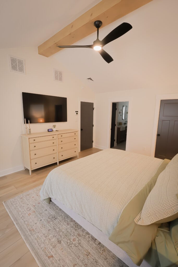 A modern bedroom with a neatly made bed, light wood dresser, large wall-mounted TV, ceiling fan, and neutral decor. The room has light wood floors, a patterned rug, and doors leading to other rooms.