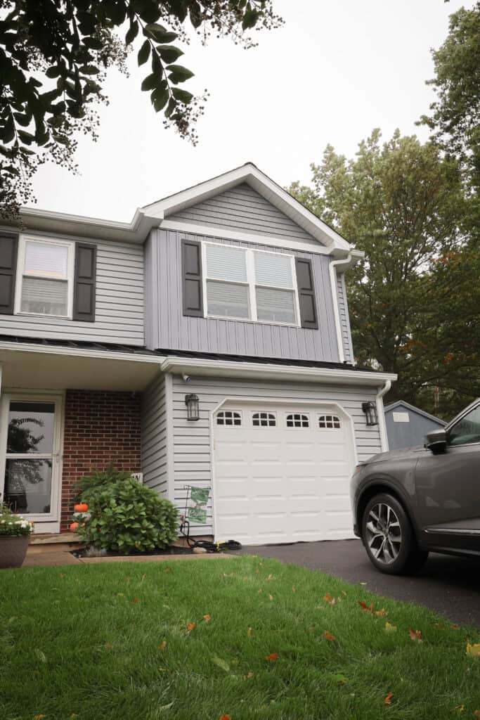A modern two-story house with gray siding, black shutters, a white garage door, and a brick wall section. A green lawn and plants are in front, and part of a gray SUV is visible in the driveway.