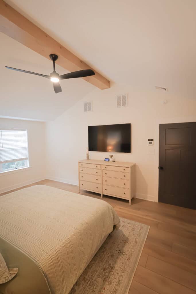 A modern bedroom with a beige bed, light wood flooring, a dresser with decor, a wall-mounted TV, a black ceiling fan, and exposed wooden ceiling beam. A window with blinds lets in natural light.