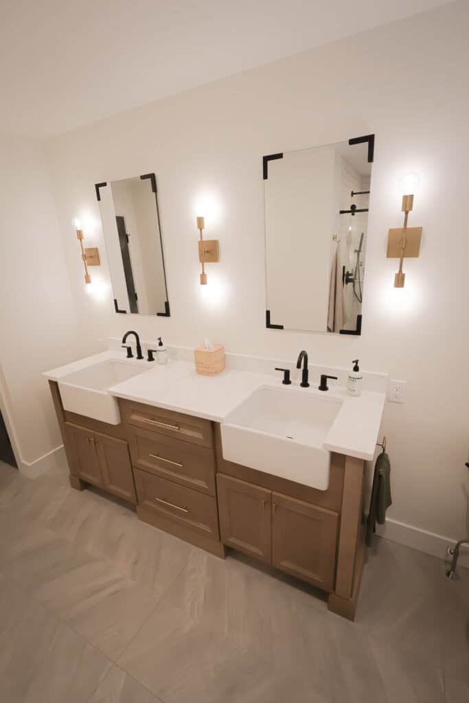 A modern bathroom with double sinks, two rectangular mirrors, black fixtures, wooden cabinetry, and wall-mounted lights beside each mirror. The countertop is white, and the floor has a herringbone tile pattern.