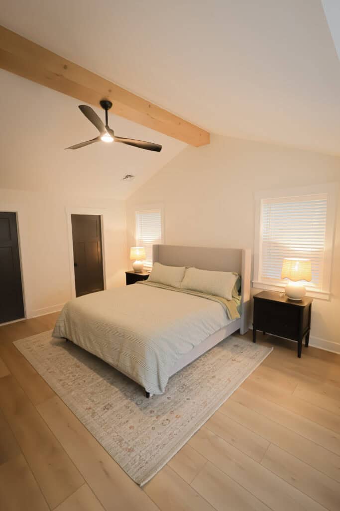 A modern bedroom with a bed centered on a light rug, two black nightstands with lamps, light wood floors, white walls, a ceiling fan, and two windows with blinds.