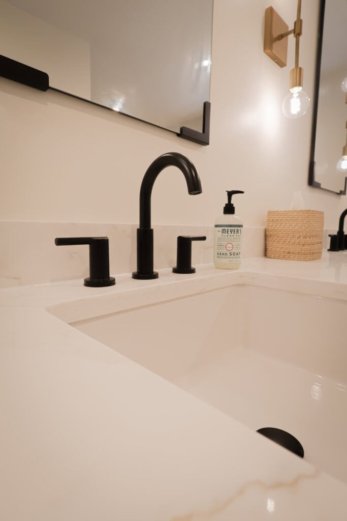 A modern bathroom sink with a matte black faucet and handles, a bottle of Meyers Clean Day hand soap, a woven container, and a wall-mounted mirror and light fixture above a white countertop.
