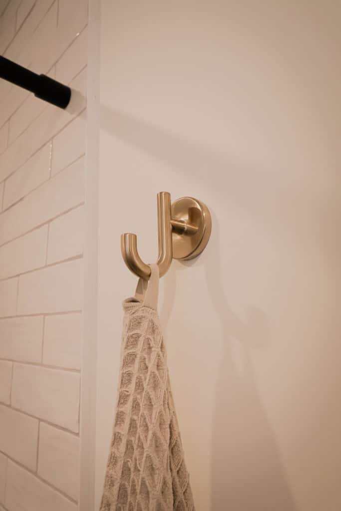 A beige textured towel hangs on a gold double wall hook mounted on a white wall next to white subway tiles.