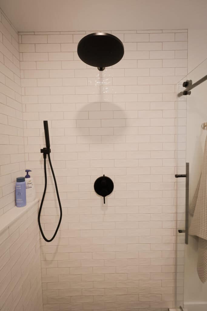 A modern shower with white subway tile walls, a round black overhead showerhead, a black handheld shower, a glass door, and bottles of soap on a built-in shelf.