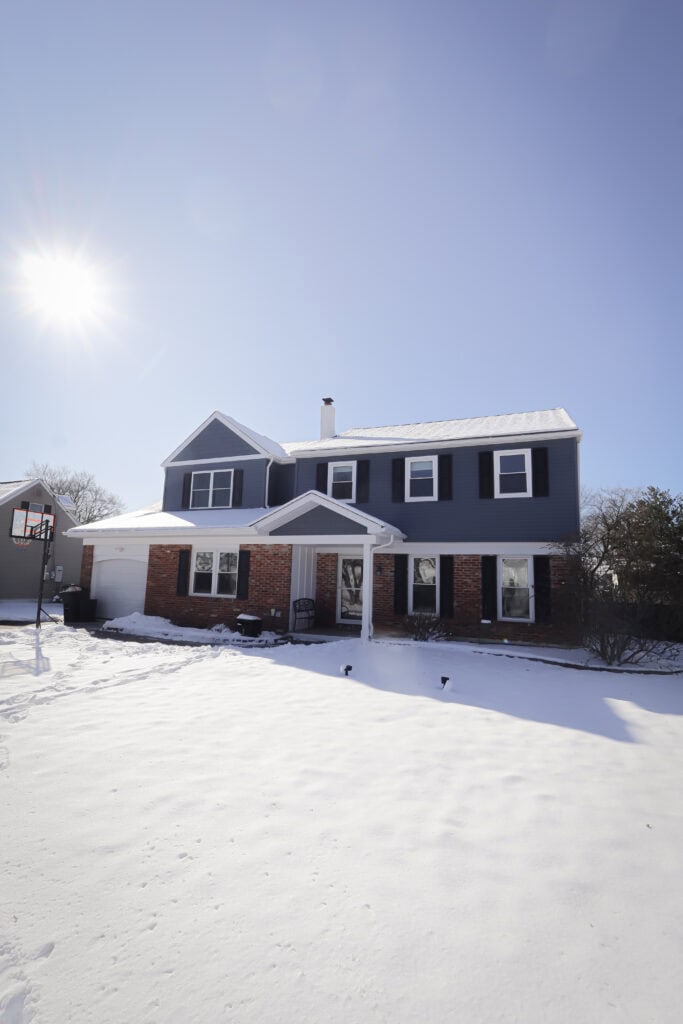 A two-story house with a dark blue exterior, white trim, and a brick lower facade sits under a clear blue sky with the sun shining, surrounded by a snow-covered yard.