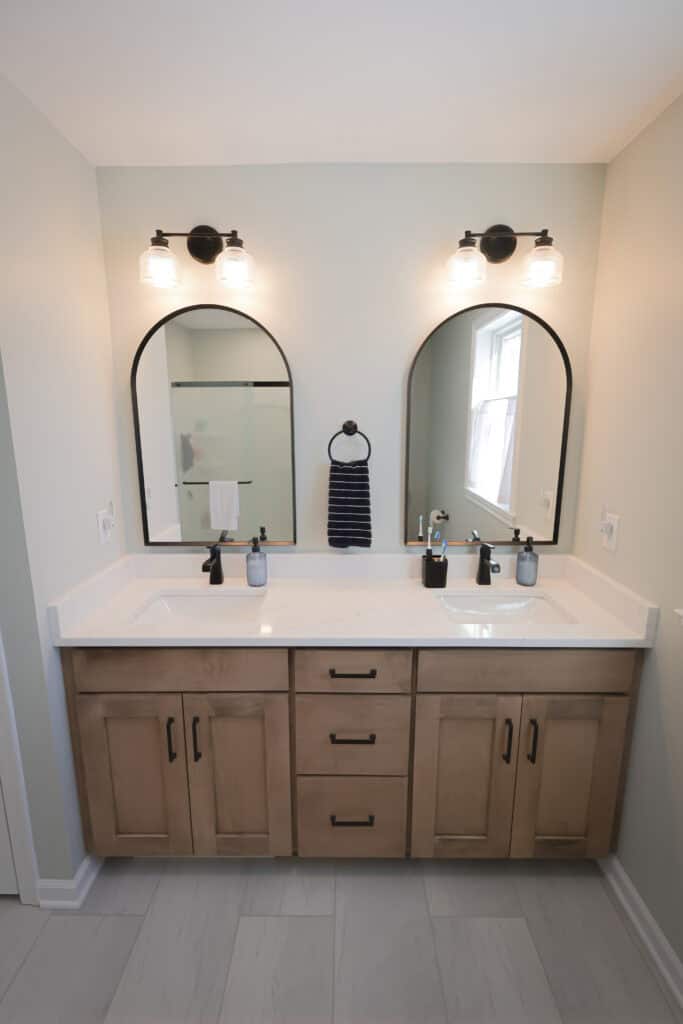 A modern bathroom with a double-sink vanity, light wood cabinets, two arched mirrors, black fixtures, wall-mounted lights, and a towel hanging between the sinks. Light gray walls and tile flooring complete the look.