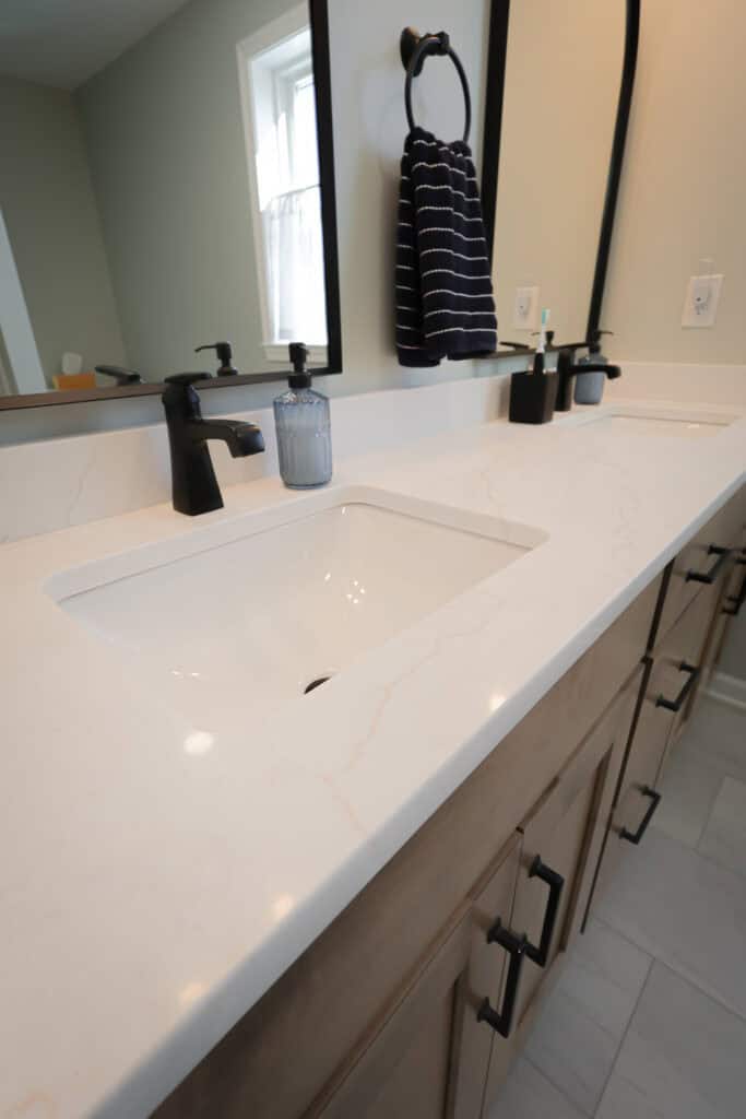 A modern bathroom vanity with a white marble countertop, rectangular sink, black faucet, soap dispenser, and a black towel ring holding a striped towel, all under a wall-mounted mirror.