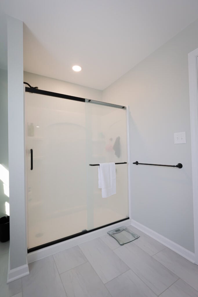 A modern bathroom with a glass shower door, black hardware, a white towel hanging on the door handle, a glass scale on the floor, and light-colored tile flooring and walls.