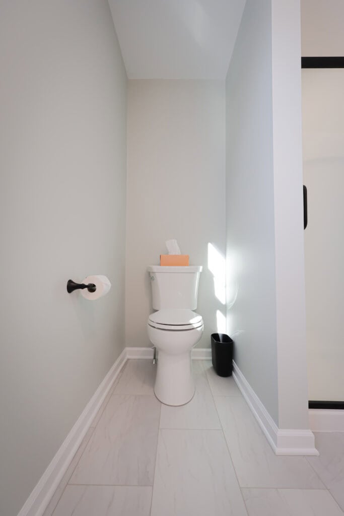 A modern bathroom with a white toilet, a wall-mounted toilet paper holder, a black trash bin, light gray walls, and white tile flooring. A box of tissues sits on the toilet tank.