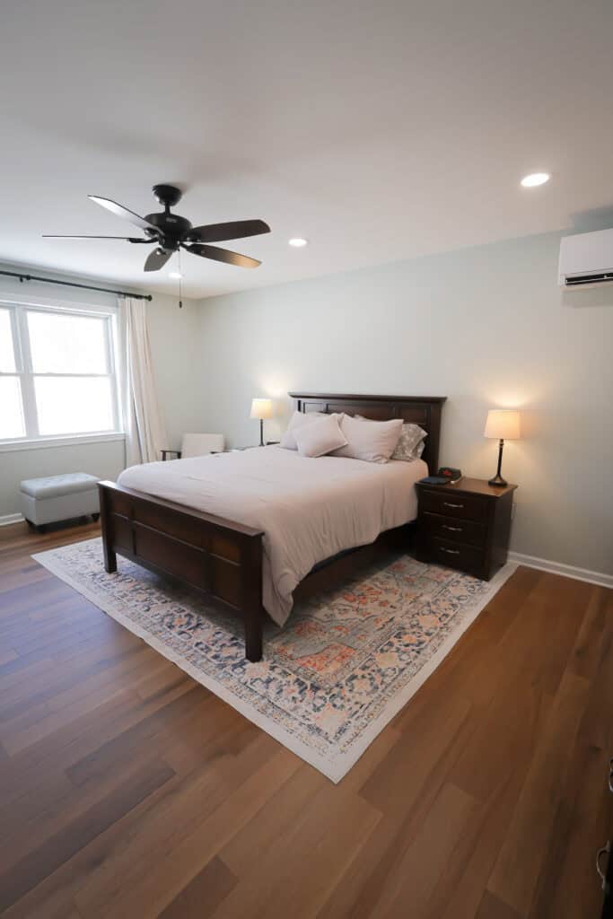 A modern bedroom with a wooden bed, beige bedding, two nightstands with lamps, a patterned rug, hardwood floor, ceiling fan, and a large window with white curtains letting in natural light.