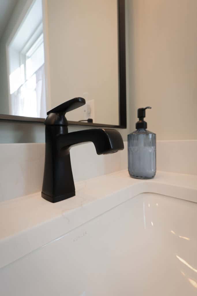 A modern bathroom sink with a matte black faucet and a translucent blue soap dispenser on a white countertop, positioned below a wall-mounted mirror. Natural light enters through a nearby window.