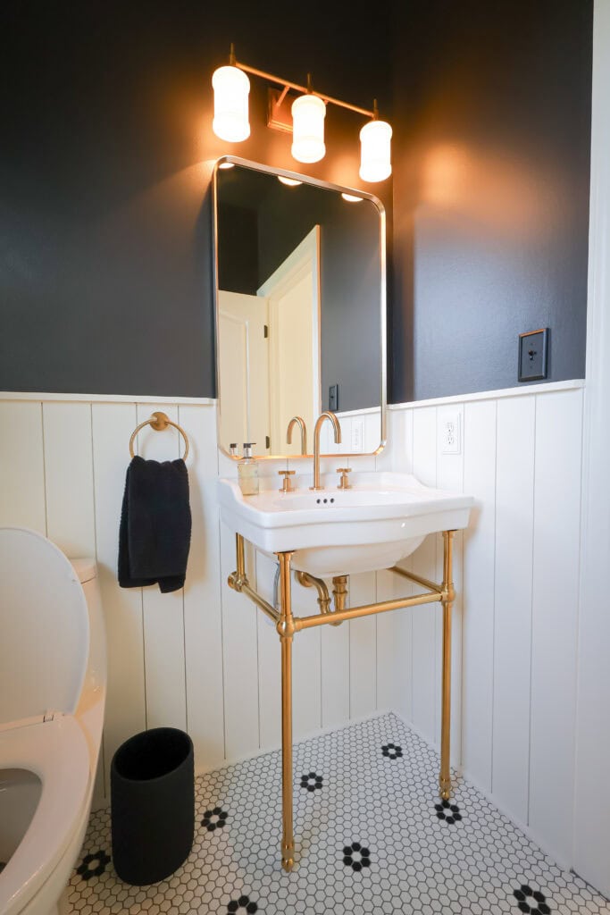 Bathroom vanity with a white wall-mounted sink, brass legs, and gold fixtures beneath a rectangular mirror and three-light vanity bar.