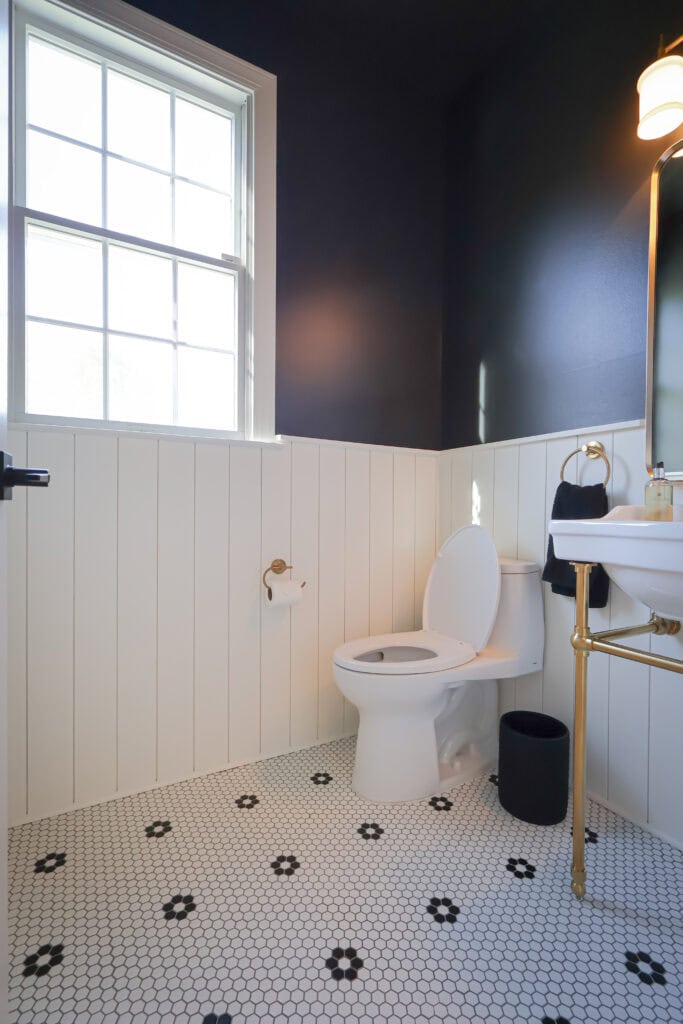 A bathroom with a toilet, pedestal sink, and white beadboard walls against dark navy paint, featuring black and white hexagonal floor tiles.