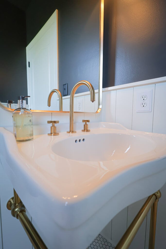 A white wall-mounted pedestal sink with a brass faucet and matching bottle of hand soap, positioned in front of a mirror and dark blue wall.