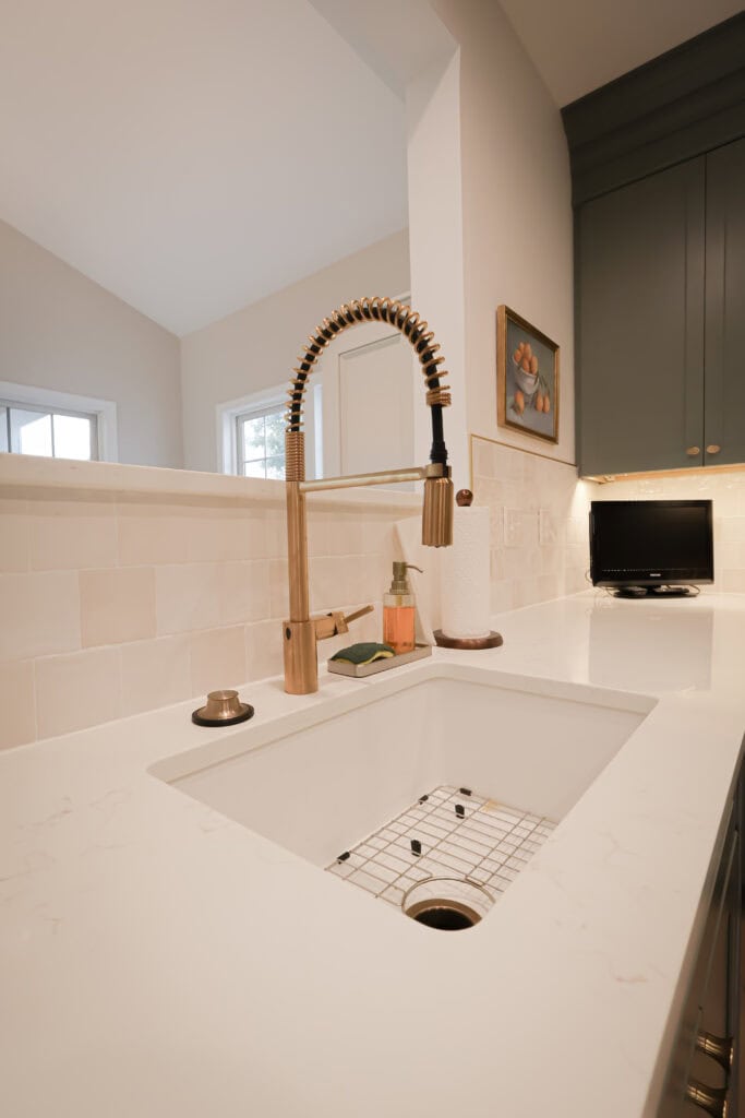 A modern kitchen sink with a gold pull-down faucet, soap dispenser, and paper towel holder on a white countertop; dark cabinets and a small screen are in the background.