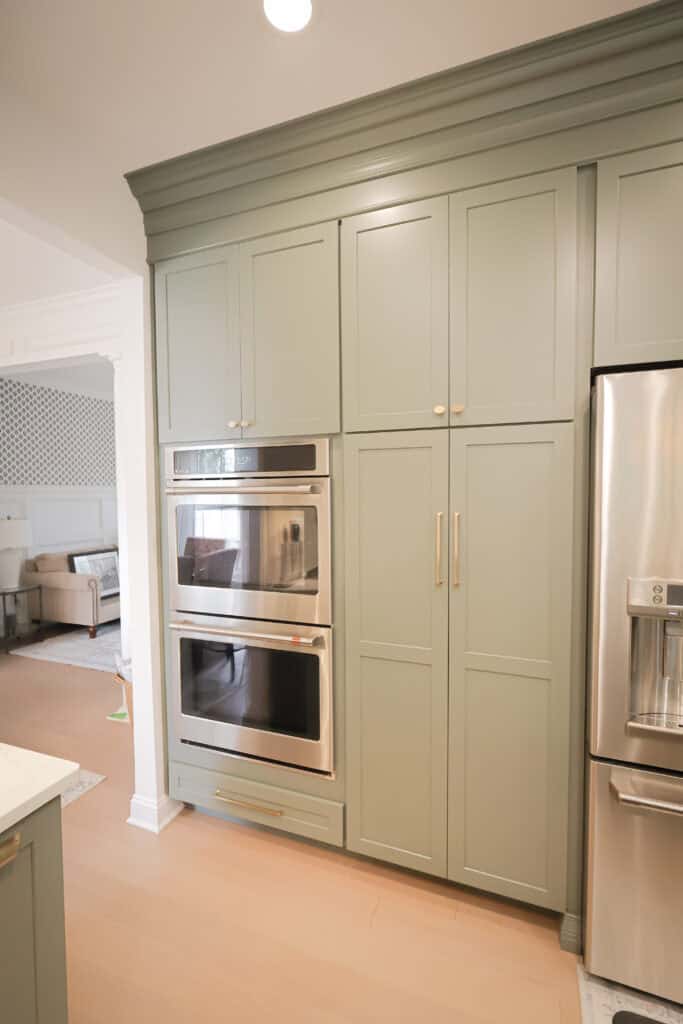 A modern kitchen features sage green cabinets with gold handles, built-in double wall ovens, and a stainless steel refrigerator with a water dispenser. A living area is visible in the background.