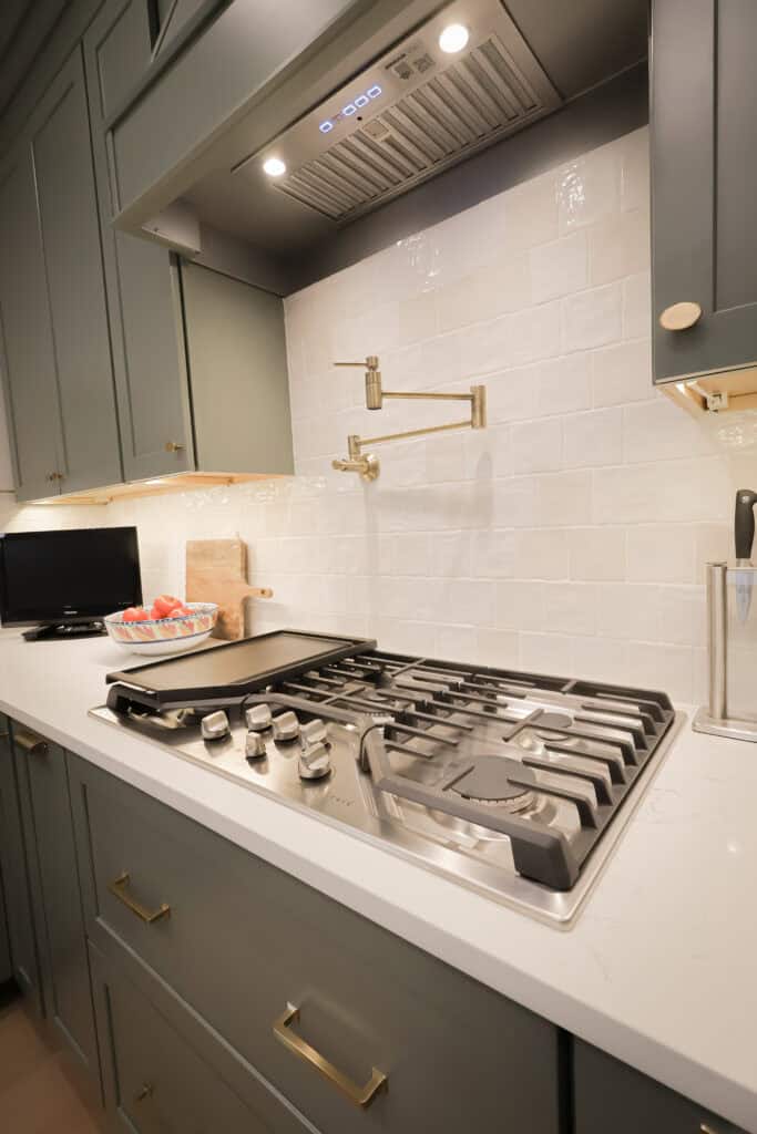 Modern kitchen with a gas cooktop, white backsplash, gray cabinets, gold handles, a wall-mounted pot filler, and a small TV on the counter beside a wooden cutting board with strawberries.