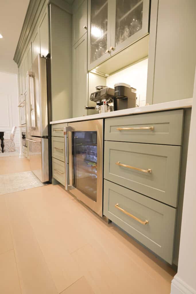 Sage green kitchen cabinets with gold handles, a built-in mini fridge with a glass door, a coffee maker, and a well-lit countertop next to a stainless steel refrigerator.