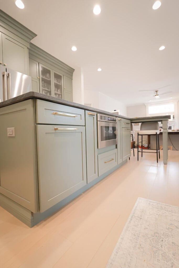 A modern kitchen features sage green cabinets and drawers, a built-in oven, light wood flooring, recessed lighting, and a central island with chairs in the background.