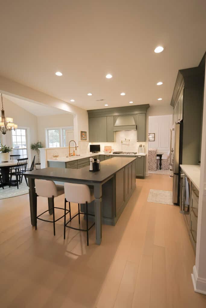 Modern kitchen with sage green cabinets, a large island with two white chairs, gold faucet, recessed lighting, light wood floors, and an adjacent dining area with a window.