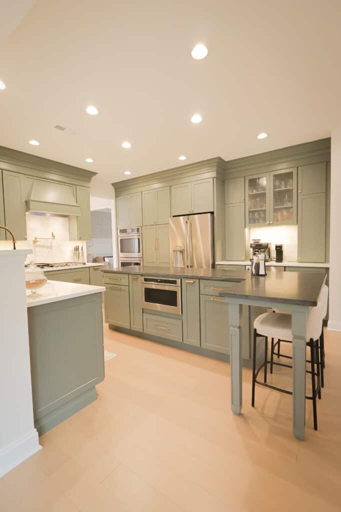 Modern kitchen with sage green cabinets, stainless steel appliances, light wood floors, white countertops, an integrated oven, and a small dining area with two white upholstered chairs. Recessed lighting brightens the space.
