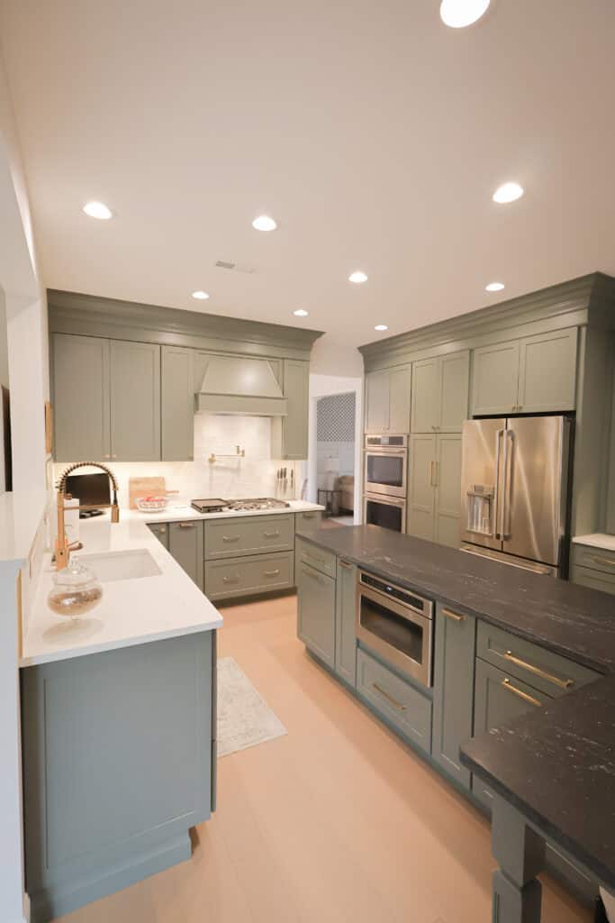 Modern kitchen with sage green cabinets, white countertops, black island, stainless steel appliances, built-in oven and microwave, and warm recessed lighting. The space is bright and tidy with a minimalist design.