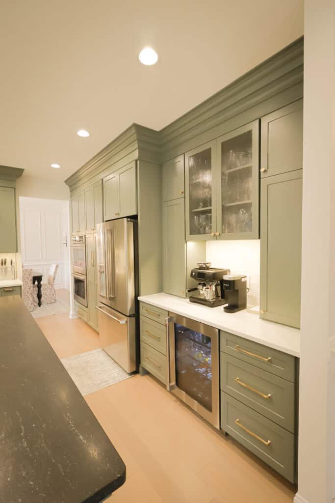 Modern kitchen with sage green cabinets, gold handles, stainless steel refrigerator, under-cabinet lighting, coffee machine, glass-front upper cabinets, wine fridge, and a black island countertop. Light wood floor and neutral walls.