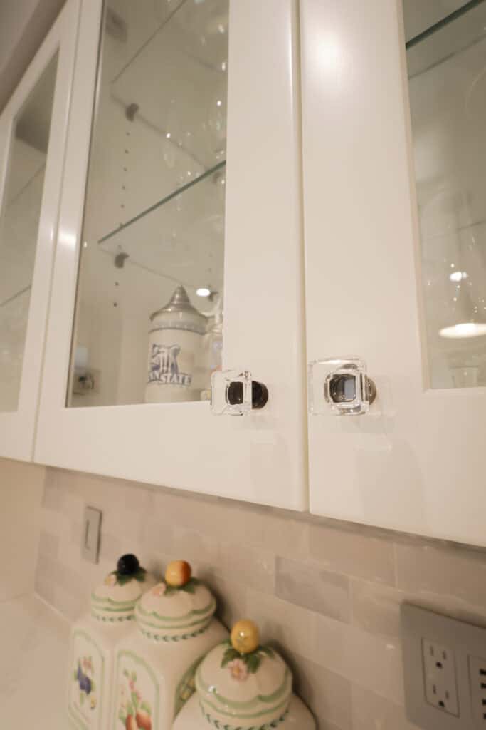 White kitchen cabinets with glass doors have clear child safety locks on the handles. Inside, glassware and jars are visible. On the counter below, decorative ceramic canisters with fruit-shaped lids are lined up.