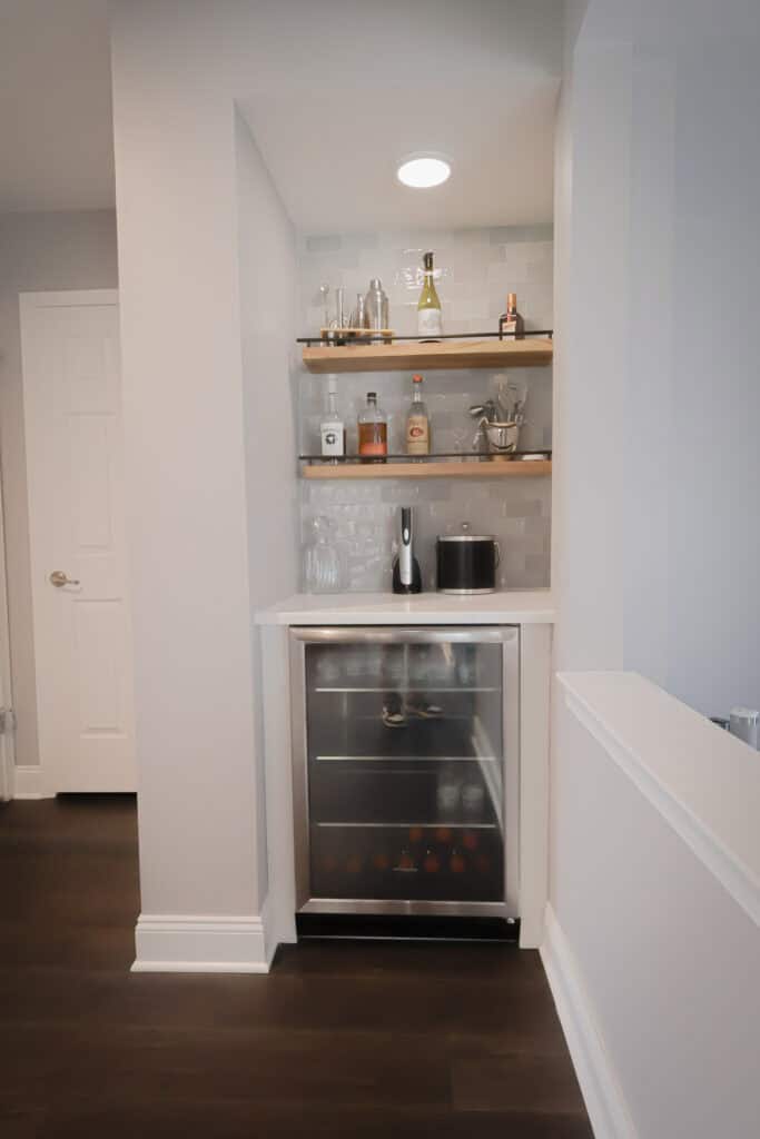 A small home bar nook with wooden shelves holding bottles and barware, a countertop with accessories, and a glass-front mini fridge stocked with drinks beneath. White tile backsplash and dark wood floor.