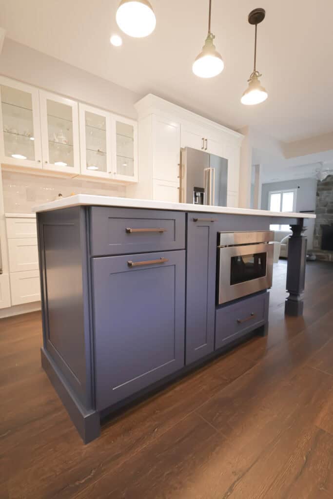 A modern kitchen with a navy blue island featuring a built-in microwave, white cabinets with glass doors, stainless steel appliances, and dark wood flooring under warm pendant lights.