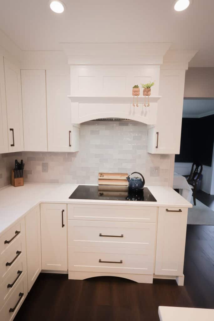 A modern kitchen with white cabinets, a white tile backsplash, a built-in stovetop with a kettle, knife block, cutting board, and two small potted plants on a white range hood shelf. Dark wood flooring is visible.
