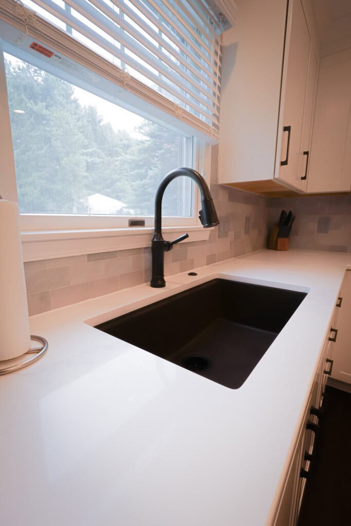 Modern kitchen countertop with a black sink and matte black faucet beneath a large window with blinds. There is a paper towel holder and a knife block on the white quartz counter, with gray tile backsplash.