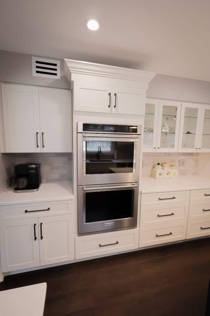 Modern kitchen with white cabinets, stainless steel double oven, glass-front upper cabinets, white countertop, coffee maker, and decorative canisters on a dark wood floor under bright lighting.