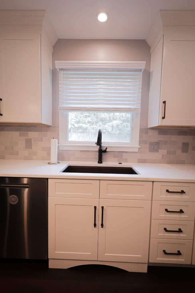 A modern kitchen with white cabinets, black handles, a stainless steel dishwasher, a black faucet over a sink, a roll of paper towels, and a window with white blinds above the sink.