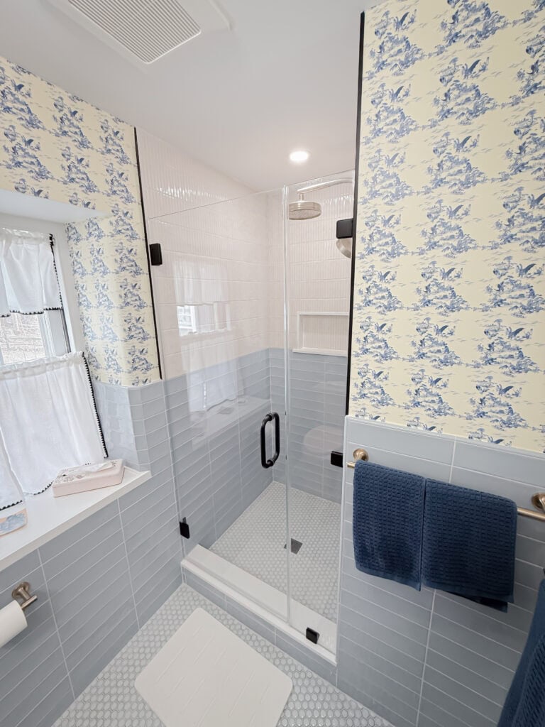 A bright bathroom with blue and white patterned wallpaper, light blue wall tiles, a glass-enclosed shower, a window with curtains, a towel rack holding blue towels, and a white bath mat on the tiled floor.