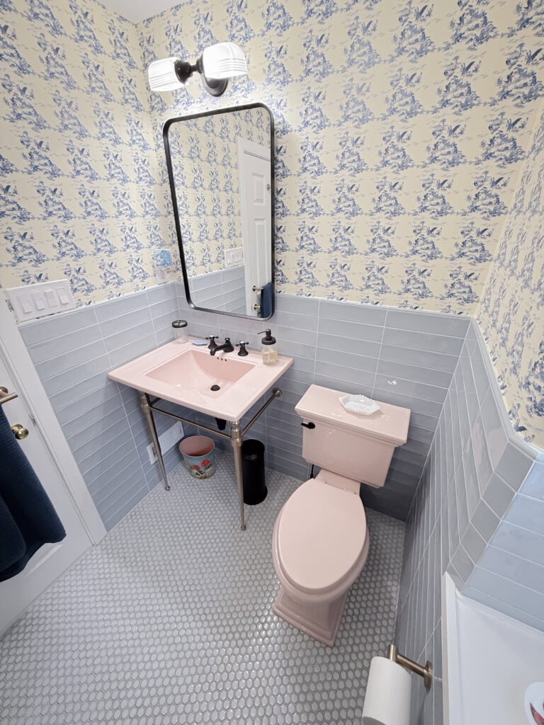 A bathroom with vintage decor, featuring a pink sink and toilet, light blue wall tiles, patterned wallpaper, a large mirror, and a white hexagon-tiled floor. A black trash bin sits below the sink.